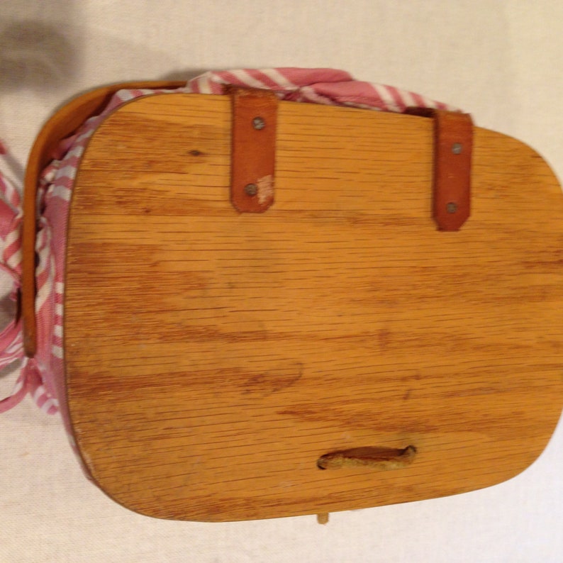 K&ouml;nnte beinhalten: Ein Vintage-Picknickkorb aus Holz mit hellbraunem Finish. Der Korb hat eine rechteckige Form mit abgerundeten Kanten und einem Ledergriff. Ein rot-wei&szlig; gestreifter Stoff ist im Inneren des Korbs sichtbar.