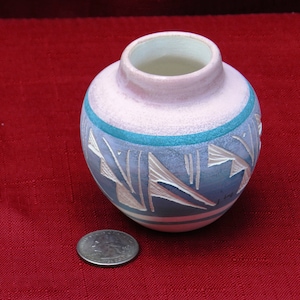 Puede incluir: Un pequeño jarrón de cerámica redondo con la parte superior rosa claro y el cuerpo azul. El jarrón presenta una banda turquesa y diseños geométricos blancos y beige. Una moneda está al lado del jarrón para la escala.