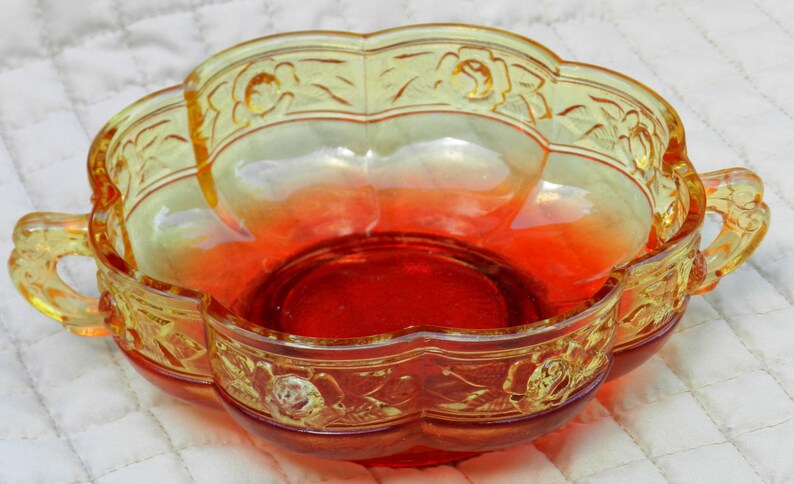 May include: A decorative, two-handled glass serving bowl with a scalloped edge. The bowl features a gradient of colors, transitioning from yellow to orange to red. Floral relief patterns adorn the rim and handles, adding a vintage touch to this kitchenware.