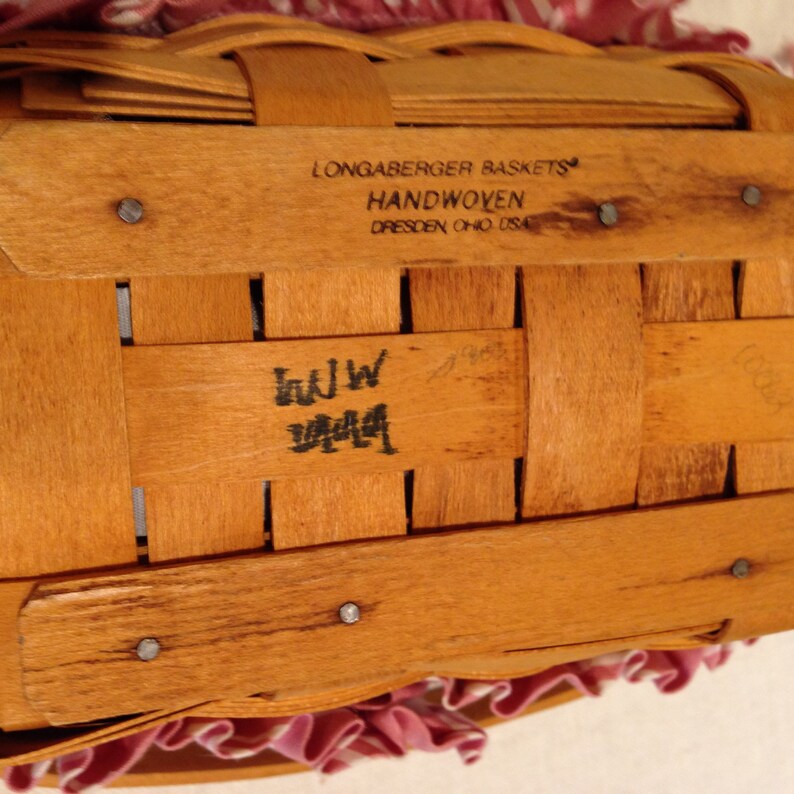 K&ouml;nnte beinhalten: Nahaufnahme eines handgewebten Longaberger-Korbs. Der Korb ist aus hellbraunem Holz gefertigt und tr&auml;gt den Text "Longaberger Baskets Handwoven Dresden, Ohio USA". Der Korb hat ein rosa-wei&szlig;es Stofffutter.