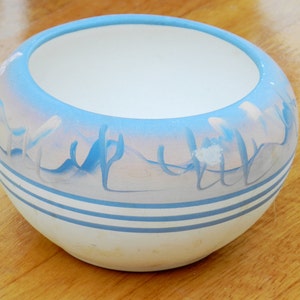 May include: A white ceramic bowl with a blue and pink glaze. The bowl has a wavy design and two blue stripes around the rim.