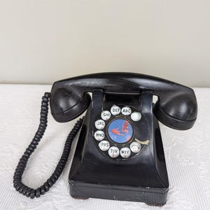 May include: A black rotary dial phone with a blue and white dial. The phone has a black cord and is sitting on a white surface.