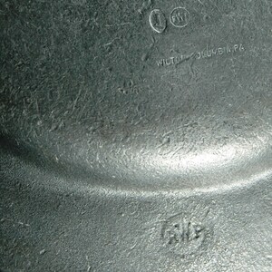 Puede incluir: Primer plano de una placa de metal gris texturizada. La placa tiene un borde ligeramente curvado y marcas visibles, incluido el texto "WILTON-COLUMBIA PA" y las iniciales "J" y "PWT".