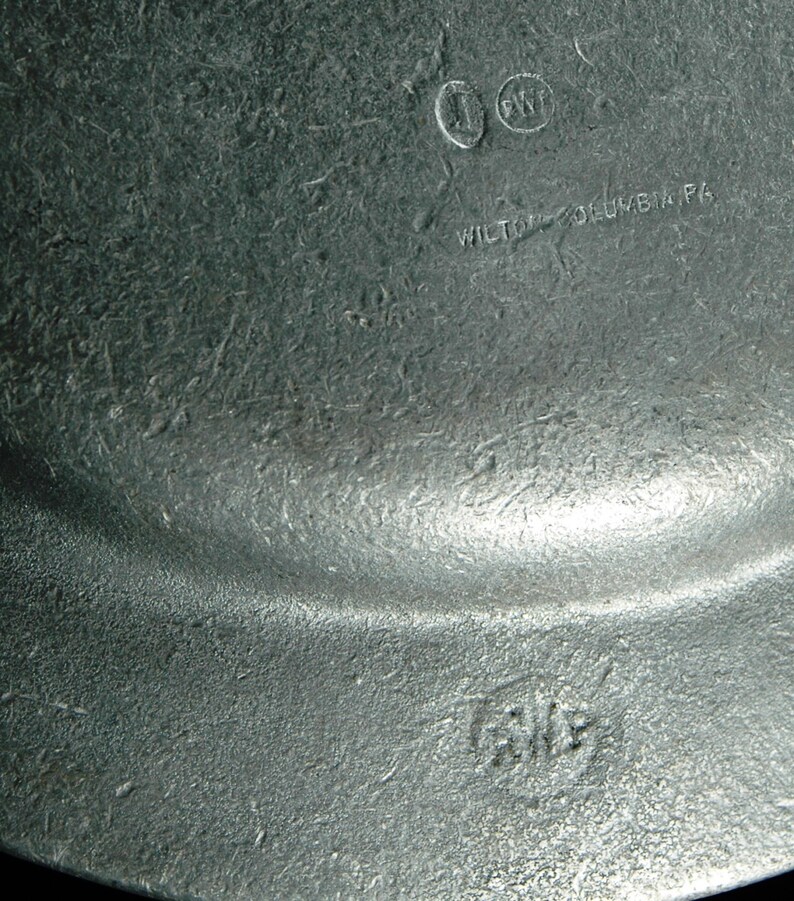 Puede incluir: Primer plano de una placa de metal gris texturizada. La placa tiene un borde ligeramente curvado y marcas visibles, incluido el texto "WILTON-COLUMBIA PA" y las iniciales "J" y "PWT".