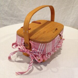 K&ouml;nnte beinhalten: Kleiner Picknickkorb aus Holz mit hellbraunem Griff und Deckel. Der Korbk&ouml;rper ist mit rosa-wei&szlig; gestreiftem Stoff mit ger&uuml;schtem Rand umwickelt. Eine passende rosa-wei&szlig; gestreifte Schleife ist an der Seite gebunden.