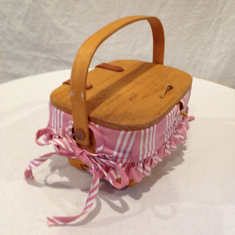 K&ouml;nnte beinhalten: Kleiner Picknickkorb aus Holz mit hellbraunem Griff und Deckel. Der Korbk&ouml;rper ist mit rosa-wei&szlig; gestreiftem Stoff mit ger&uuml;schtem Rand umwickelt. Eine passende rosa-wei&szlig; gestreifte Schleife ist an der Seite gebunden.