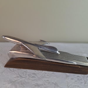 May include: A vintage silver-toned hood ornament in the shape of a streamlined airplane, mounted on a rectangular wooden base. The ornament has a sleek, aerodynamic design with a prominent wing and tail. The base is a dark brown color.