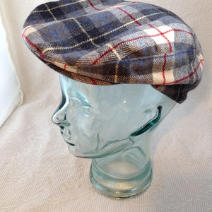 Op de afbeelding: Een geruite newsboy-pet met een grijs, blauw, wit en rood patroon. De pet is gemaakt van een zachte, textuurrijke stof.