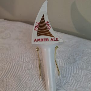 May include: White beer tap handle with a sail-shaped top. The top has the words "FULL SAIL AMBER ALE" in red lettering. The sail design is brown with brown horizontal lines. The handle has gold-colored metal accents.
