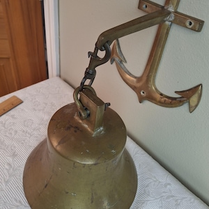 May include: A large, weathered brass ship bell suspended by a metal clip from a wall-mounted brass anchor bracket. The bell features a wide, flared base and a sturdy top attachment point, while the anchor bracket includes pre-drilled holes for mounting.