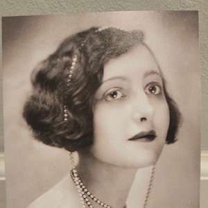 May include: A black and white photograph of a woman with dark hair styled in a bob. She wears a pearl necklace and earrings. The photograph is displayed on a small wooden easel.