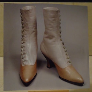 Puede incluir: Un par de botas de cuero beige claro con cierres de botón en los lados. Las botas tienen una punta puntiaguda y un tacón bajo.