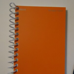 Puede incluir: Una libreta de espiral naranja con la palabra "WOCONO" impresa en la portada, y un bolígrafo azul con un capuchón blanco y un logotipo en el capuchón.