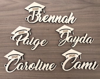 Może przedstawiać: Drewniane wycinanki z imionami absolwentów z czapką absolwenta i rokiem 2020. Imiona to Brennah, Paige, Jayda, Caroline i Cami. Wycinanki są z jasnego drewna, a tło jest z drewna o średnim odcieniu.