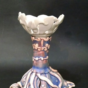 May include: A white ceramic candle holder with a floral design. The base is blue and purple with a swirling pattern. The holder has a wide, open top.