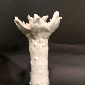 May include: White ceramic vase with a unique, organic design. The vase has a wide, flared opening and a tall, slender body. The base of the vase is shaped like a tree trunk with four roots.