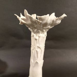 May include: A white ceramic candle holder with a unique, organic design. The base features three curved feet, supporting a tall, textured stem that culminates in a flower-like top. The overall height is approximately 10 inches.