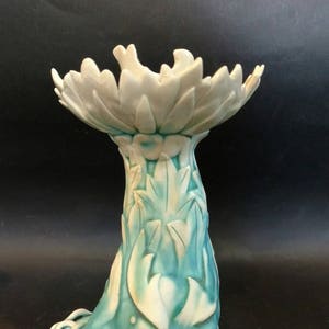 May include: A ceramic decorative pedestal in shades of teal and white. The pedestal features a floral design at the top and a textured stem with leaf patterns. The base has a unique, curled foot design. The overall aesthetic is artistic and decorative.