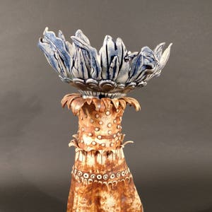 May include: A ceramic hand-shaped sculpture with a blue and white flower-shaped bowl on top. The sculpture is decorated with brown and white glaze and has a textured surface.