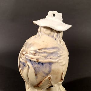 May include: A white ceramic sculpture of a bird with a white hat. The bird has a round body with blue and brown accents. The bird is standing on a base with a white glaze.
