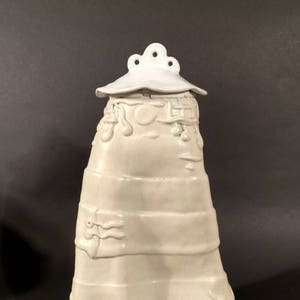 May include: White ceramic jar with a lid and intricate raised designs. The jar has a rounded base and a tall, slender body. The lid has a scalloped edge and a small knob on top.