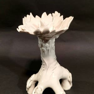 May include: White ceramic vase with a floral design. The vase has a wide, open top and a base with three legs that resemble stylized tree roots.