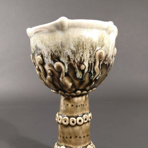 May include: A ceramic goblet with a brown and white glaze. The goblet has a wide, round bowl and a tall, slender stem. The bowl is decorated with raised, swirling designs. The stem is decorated with a band of small, round dots.
