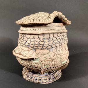 May include: A ceramic jar with a lid, decorated with intricate patterns and a textured surface. The jar has a light brown color with hints of green and purple.