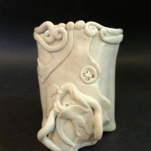 May include: White ceramic vase with a textured, organic design. The vase has a wide mouth and a narrow base. The design features a series of raised lines and curves that create a flowing, abstract pattern.