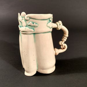 May include: White ceramic mug with a teal glaze and a rope-like handle. The mug has a decorative design of clouds around the top.