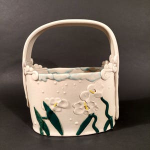 May include: White ceramic planter with a handle, decorated with green leaves and white flowers. The planter has a textured surface with small white dots.