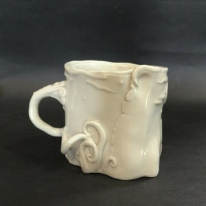 May include: A white ceramic mug with an organic, textured design. The mug features a curved handle and decorative, swirling relief patterns on the exterior. The overall shape is irregular, giving it a handcrafted appearance. The mug is suitable for hot or cold beverages.