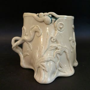 May include: A white ceramic vase with a leafy design and a green glaze around the rim. The vase has a unique, organic shape and a textured surface.