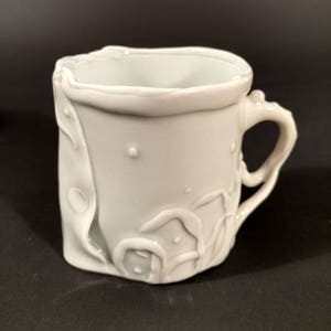 May include: White ceramic mug with a unique, textured, and slightly irregular design. The mug has a handle and a rounded base.