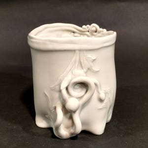 May include: A white ceramic vase with a unique, organic shape and intricate floral designs. The vase has a wide opening and a rounded base.