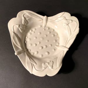 May include: A white ceramic dish with a raised design of leaves and flowers. The center of the dish has a textured surface with small circles.