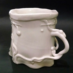 May include: White ceramic mug with a unique, organic, and flowing design. The handle is sculpted to resemble a vine or branch. The mug has a smooth, glossy finish.