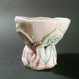 May include: A white ceramic vase with a unique, organic design. The vase has a wide, open top and a narrow base. The surface is decorated with raised, textured details in shades of green and pink.