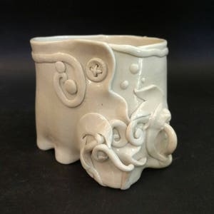 May include: A white ceramic cup with a unique, textured design. The cup has a rounded base and a wide, open top. The surface of the cup is decorated with raised, swirling patterns and a button-like detail.