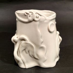 May include: White ceramic vase with a textured, organic design. The vase has a wide, round opening and a narrow base. The design features raised, flowing lines and curves, resembling leaves and vines.