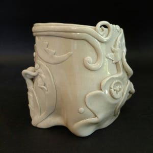 May include: A white ceramic vase with a unique, organic design. The vase has a textured surface with raised floral patterns and a wavy, uneven base.