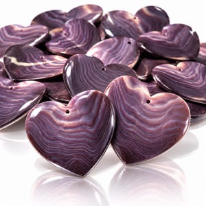 Könnte beinhalten: Eine Sammlung herzförmiger Anhänger aus poliertem violettem Muschelmaterial. Jedes Stück weist ein ausgeprägtes natürliches, wellenförmiges Maserungsmuster in tiefen Violett- und Weißtönen auf, mit einem kleinen Loch im oberen Bereich.