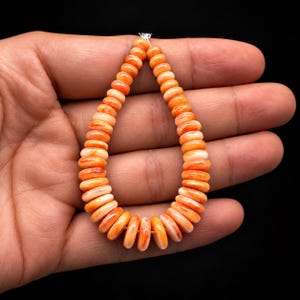May include: A strand of round, flat, disc-shaped beads in varying shades of orange and white. The beads are arranged in a teardrop shape, featuring a smooth, polished surface with natural color variations throughout each individual bead piece.