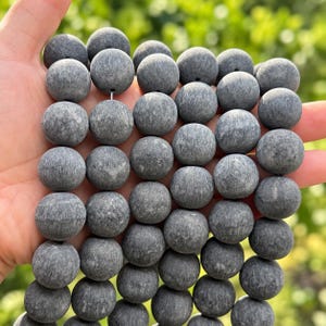 May include: A collection of round, dark gray beads with a matte, textured finish. Each bead features a subtle, fibrous pattern across its surface and a small, centered hole for stringing. The beads are arranged in multiple vertical rows.