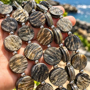 May include: A multi-strand necklace featuring circular, flat, brown and black patterned stone beads with a textured, marbled appearance. The beads are separated by small, polished, spherical silver-tone metal spacers, creating a repeating pattern.
