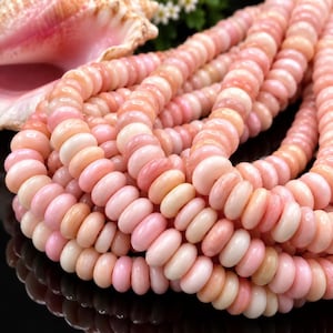 May include: A strand of smooth, round, light pink beads with a glossy finish. The beads are arranged in multiple rows, showcasing a consistent pale pink color palette. A large, textured, light pink seashell is visible in the upper left corner.