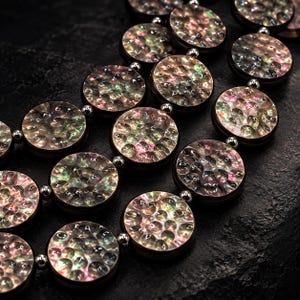 May include: A necklace featuring multiple circular discs with a hammered, iridescent finish that displays shifting hues of pink, green, and gold. The discs are connected by small, polished silver-toned metal beads in a multi-strand arrangement.
