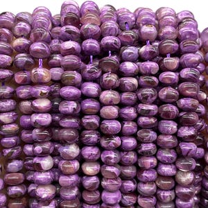 May include: Close-up of a strand of purple charoite gemstone beads. The beads are barrel-shaped, with a mix of light and dark purple hues and some brown inclusions. The beads are strung together with a purple cord, ready for jewelry making.