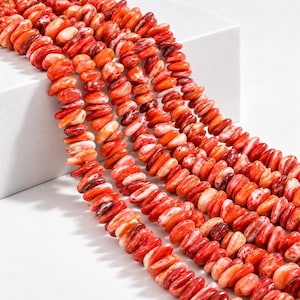 May include: Multiple strands of small, irregular, flattened beads in vibrant shades of orange, red, and white. The beads have a polished, marbled appearance with swirling color patterns, arranged in parallel lines that drape across the frame.