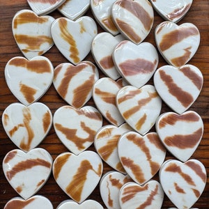 May include: A collection of heart-shaped objects featuring a smooth, off-white base color with irregular, horizontal brown streaks and patterns across the surface. Each heart has a glossy finish and is arranged in a loose, overlapping cluster.